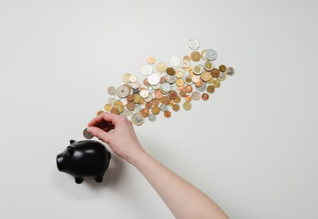Photograph of a black piggy bank surrounded by cons with someone's hand dropping a coin in